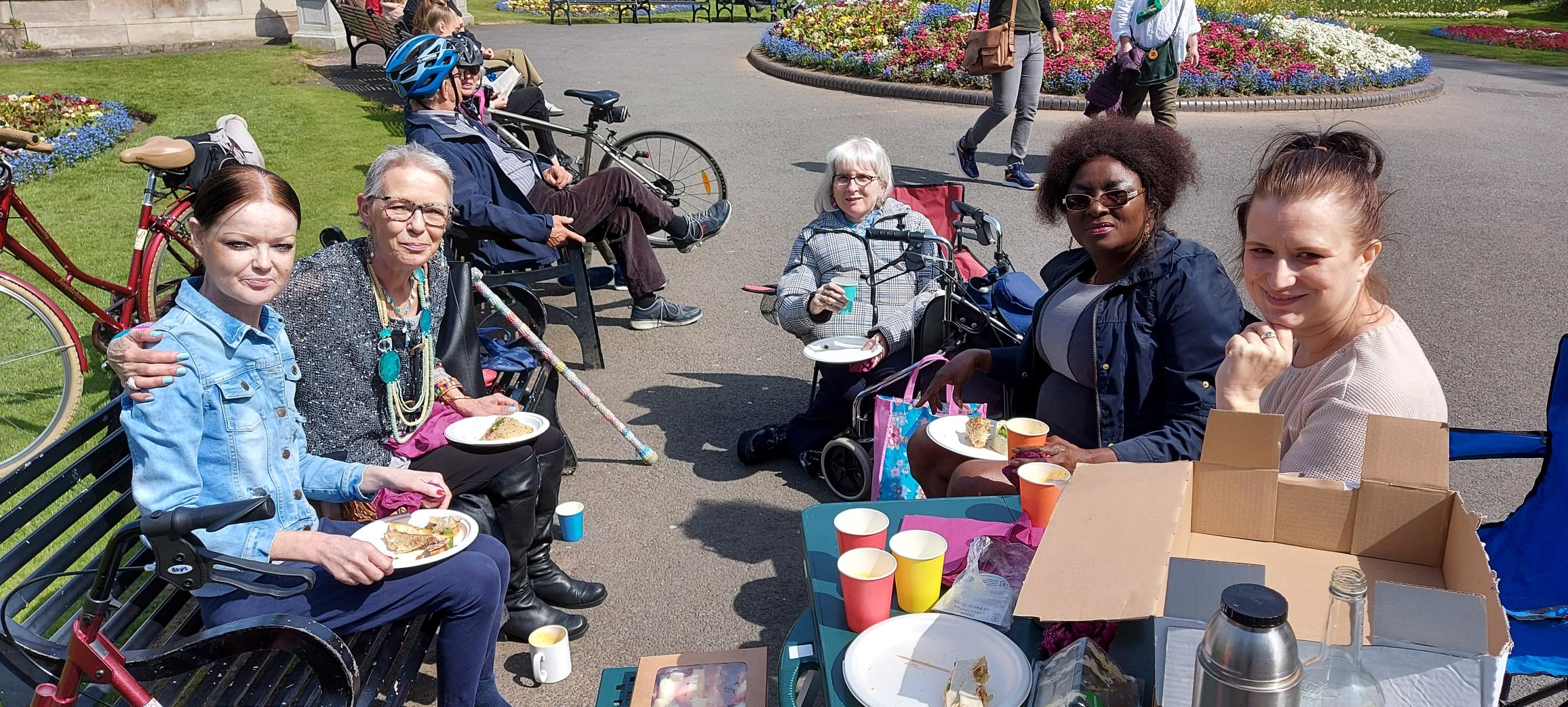 This photograph shows a group of five women looking at the camera while they enjoy a picnic lunch in a park.  These are volunteers and members of the Women’s Group with Community Navigator Clare Bennett.