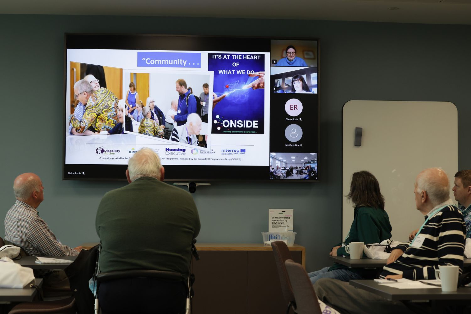 This photograph shows a group of people in the room together looking at a large screen which includes a number of people who are joining the even via Zoom.  This is to show how the project has continued to adapt to the covid pandemic by using technology to facilitate hybrid meeting.
